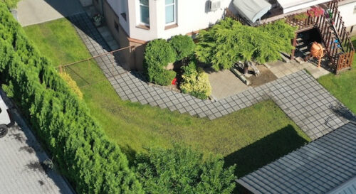 Aerial view of a house corner with a lush garden. Pathways meander through green grass and dense bushes, creating a serene and inviting atmosphere.